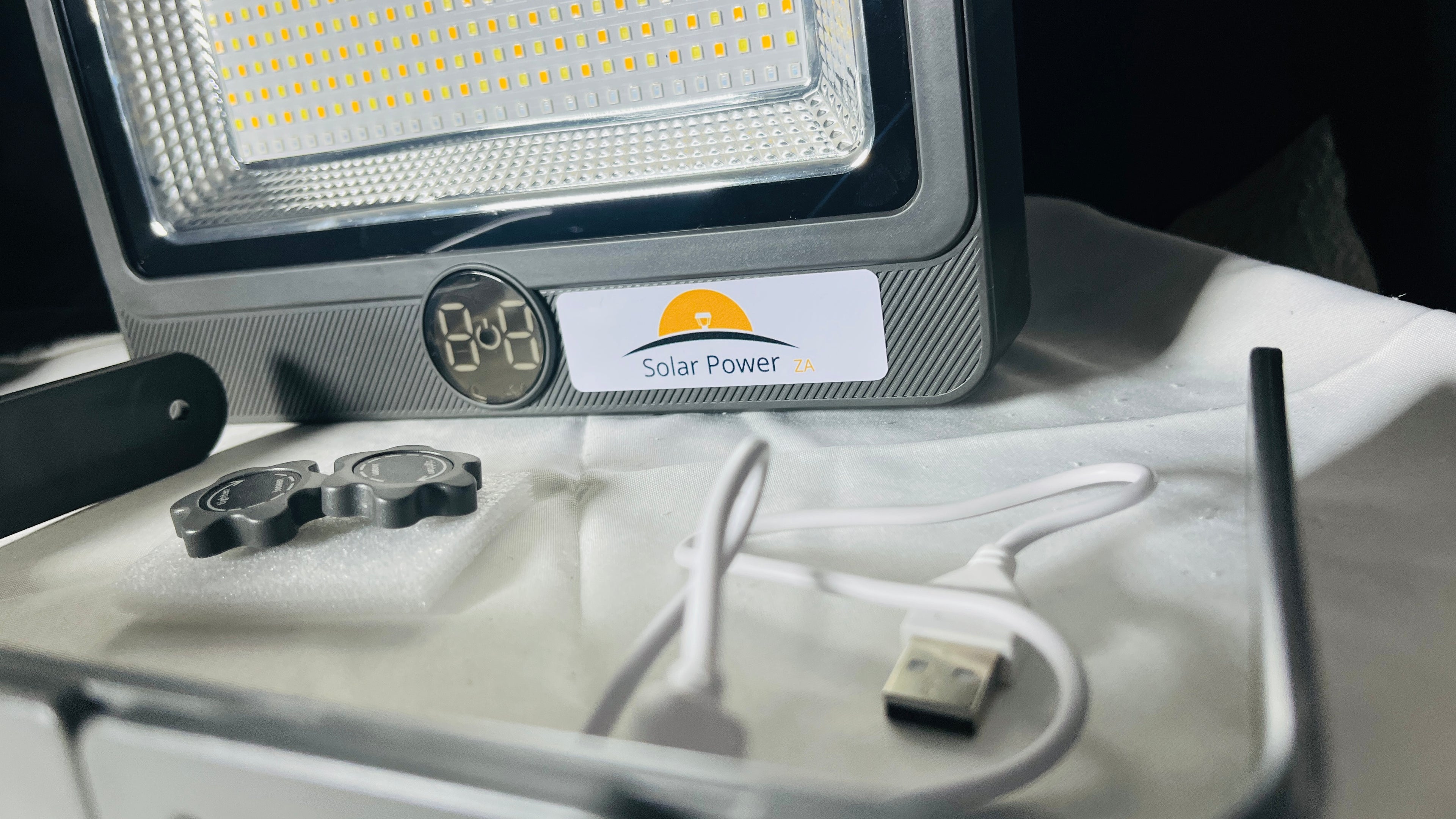 A solar-powered LED light panel with a digital display and USB charging cable, shown with mounting hardware on a white surface.