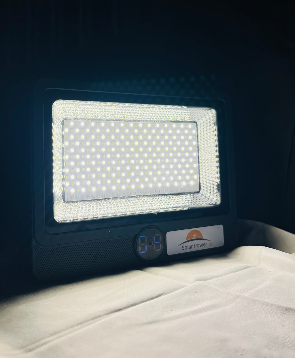 A solar-powered LED light panel with a black housing, featuring a bright illuminated white LED grid and a small digital display showing '8.4' with a 'Solar Power' label, resting on a white fabric surface against a dark background.